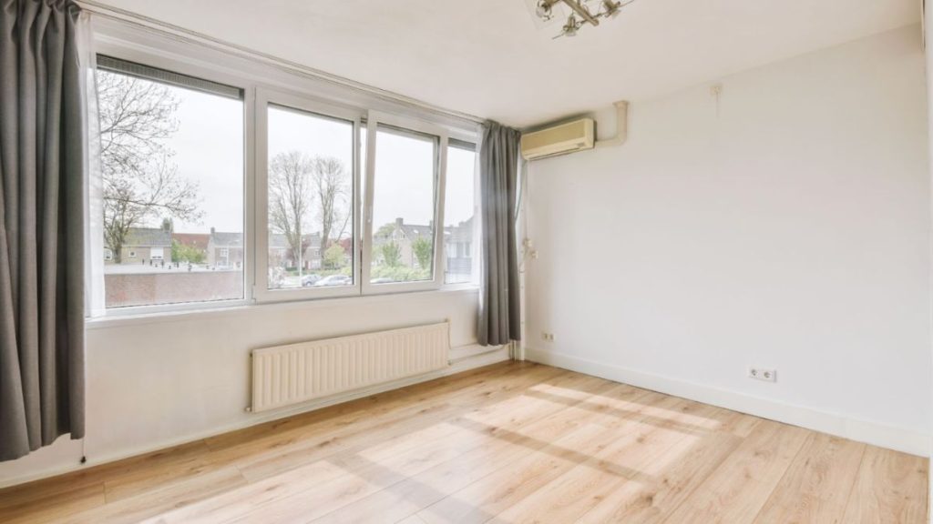 A vacant room featuring a window and polished wooden floors, illuminated by natural light streaming through the glass.