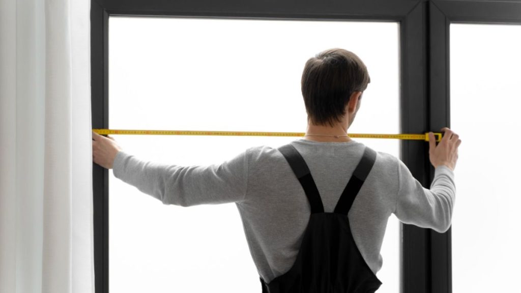 A man carefully measures a window frame using a tape measure to ensure accurate dimensions for installation.