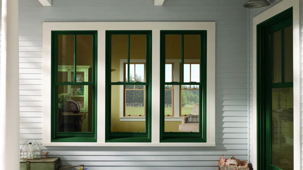 A cozy front porch features three 400 Series double-hung windows with dark green frames and white trim set against light gray siding. Inside, yellow walls and wooden furniture create a warm and inviting space.