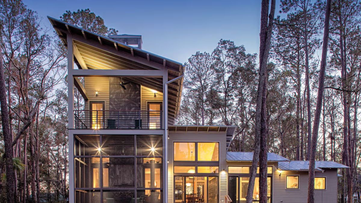 A modern two-story house nestled among tall trees, showcasing a large picture window and patio doors on the ground floor. The exterior blends wood and metal siding, with a screened-in porch below and an open balcony above. Warm light spills through the windows, offering a cozy contrast against the twilight sky.
